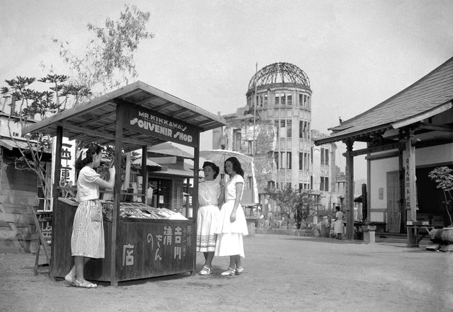 Pada 3 Agustus 1951, yaitu enam tahun setelah bom atom dijatuhkan tepat di atas titik ini di Hiroshima, sebuah kios cenderamata berdiri di jalan dekat kubah Balai Industri yang hancur. Kios ini dioperasikan oleh Kiyoshi Yoshikawa, yang juga terluka dalam peristiwa pengeboman tersebut. (Photo by AP Photo/Kyodo via The Atlantic)